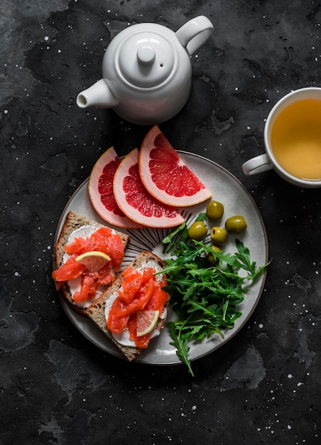 Delicioso desayuno salmón crema queso grano pan sándwiches rúcula aceitunas rodajas de pomelo y té verde sobre un fondo oscuro vista superior