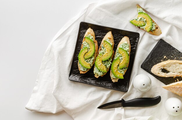 Delicioso desayuno saludable con sándwiches abiertos de baguette, ricotta, espinacas y aguacate en un plato negro sobre la mesa blanca. copia espacio