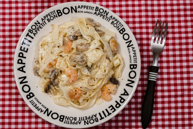 Delicioso fettuccine com molho de creme de cogumelos e camarão em um prato que diz bon appetite com toalha de mesa quadriculada vermelha. cozinha italiana.