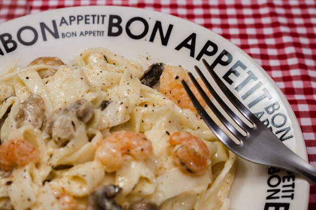 Delicioso fettuccine com molho de creme de cogumelos e camarão em um prato que diz Bon Appetite com toalha de mesa quadriculada vermelha. Cozinha italiana.