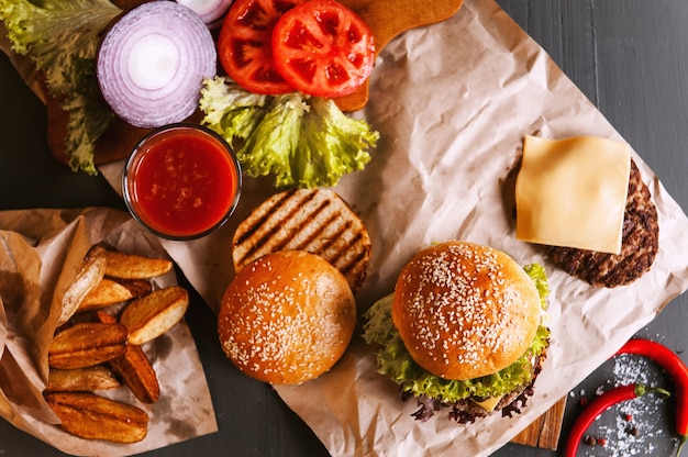 Delicioso hambúrguer caseiro fresco em uma mesa de madeira. próximo ao componente para hambúrguer, bandejas de madeira, batatas fritas e pimenta. Um copo de suco de tomate