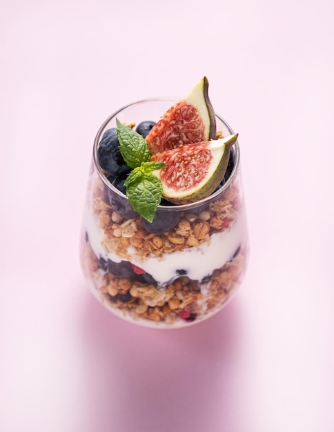 Delicioso iogurte natural com granola figos e bagas em um copo em um fundo rosa