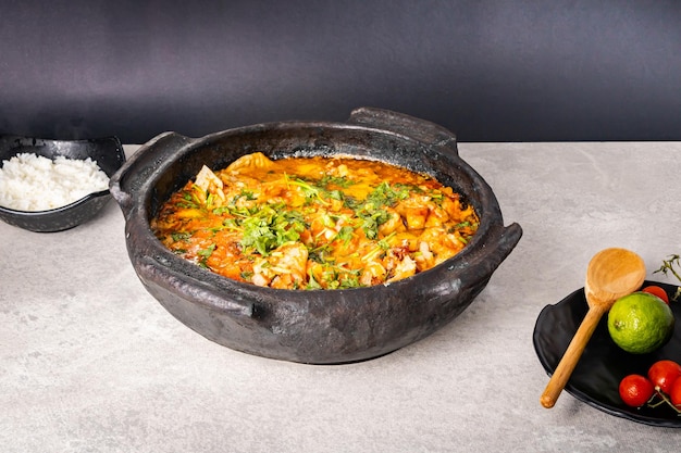 Delicioso moqueca de pescado brasileño con tomate, cebolla, aceite de oliva, cilantro y semilla de orocum Hecho en olla de barro sobre mesa blanca y fondo negro