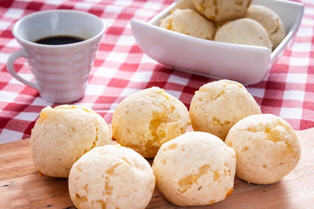 Foto delicioso pão de queijo um prato típico brasileiro em uma tábua de madeira e uma toalha de mesa quadriculada ao lado de uma xícara de café