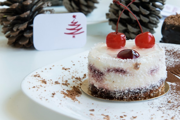 Delicioso pastel de coco / cereza de invierno con una tarjeta de felicitación sobre fondo blanco y cono de pino en el fondo