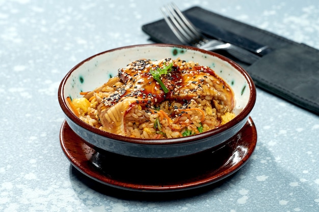 Delicioso plato panasiático: arroz wok con anguila, verduras y salsa agridulce en un recipiente verde sobre una superficie gris
