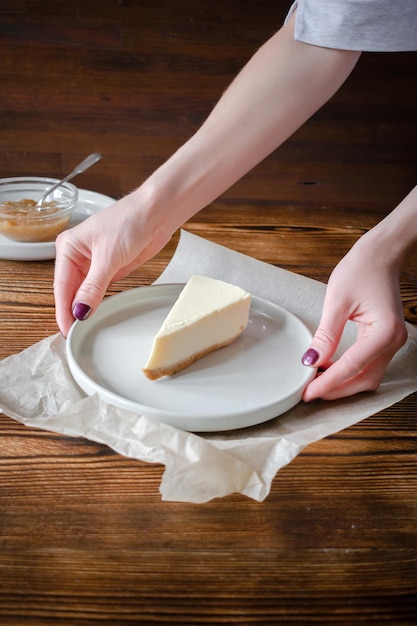 Delicioso postre dulce clásico cheesecake Nueva York sobre fondo rústico de madera Manos de mujer sirviendo rebanada de pastel sabroso en plato de cerámica con tenedor de postre y mermelada de manzana