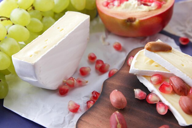 Delicioso queso sobre tabla de madera y papel pergamino de cerca Camembert con semillas de granada, uvas verdes y nueces sobre un fondo azul.