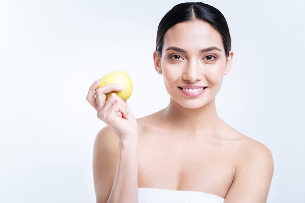 Delicioso refrigerio. Encantadora mujer joven alegre sosteniendo una manzana amarilla en su mano mientras está de pie envuelto en una toalla, posando contra una pared blanca