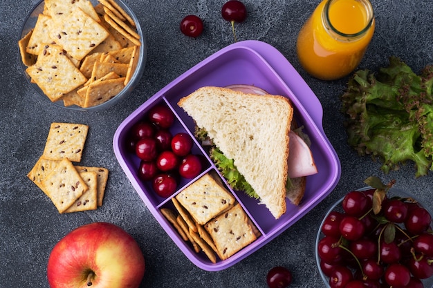 Delicioso sanduíche saudável em uma lancheira, biscoitos e cerejas. Leve o almoço com você para a escola ou para o escritório. Suco em uma garrafa e uma maçã.