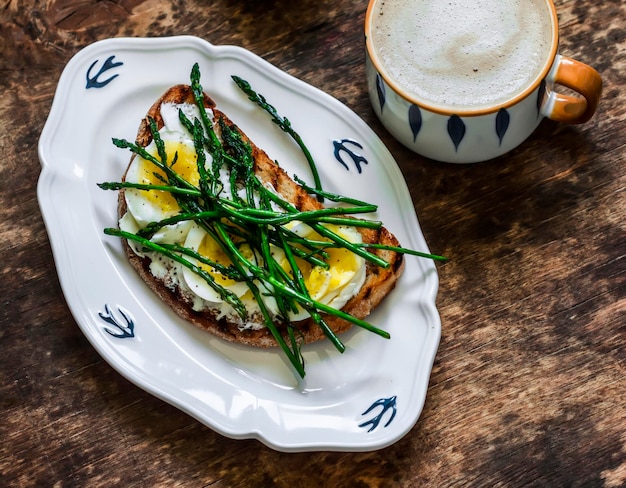 Delicioso sándwich de desayuno con queso crema, huevo hervido, espárragos jóvenes y capuchino en una vista superior de fondo de madera