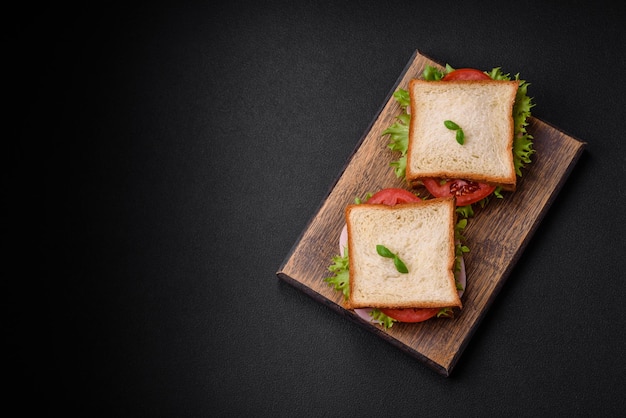 Delicioso sándwich con tostadas de jamón, tomates, queso y lechuga con sal, especias y hierbas sobre un fondo de hormigón oscuro