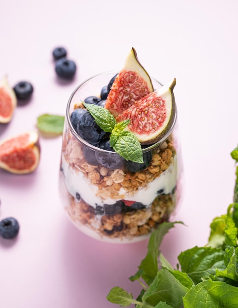 Delicioso yogur natural con higos de granola y arándanos en un vaso sobre un fondo rosa con menta