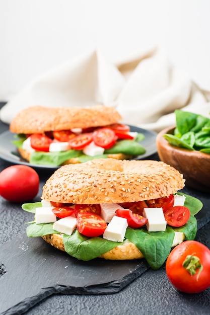 Deliciosos bagels prontos para comer recheados com tomate, queijo feta e folhas de espinafre em uma placa de ardósia sobre um fundo preto. Lanche leve e saudável. Visão vertical