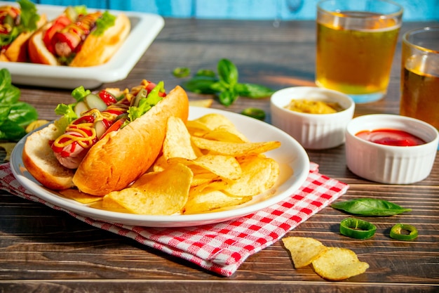 Deliciosos cachorros-quentes caseiros com batatas fritas e copos de cerveja na mesa de madeira