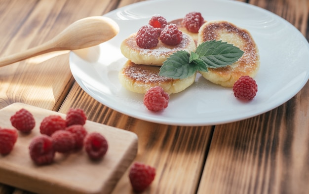 Deliciosos cheesecakes ou panquecas com açúcar de confeiteiro na chapa branca na mesa de madeira com framboesas e folha de hortelã