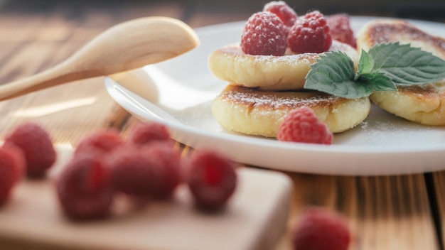 Deliciosos cheesecakes ou panquecas com açúcar em pó na chapa branca na mesa de madeira com framboesas e folha de hortelã