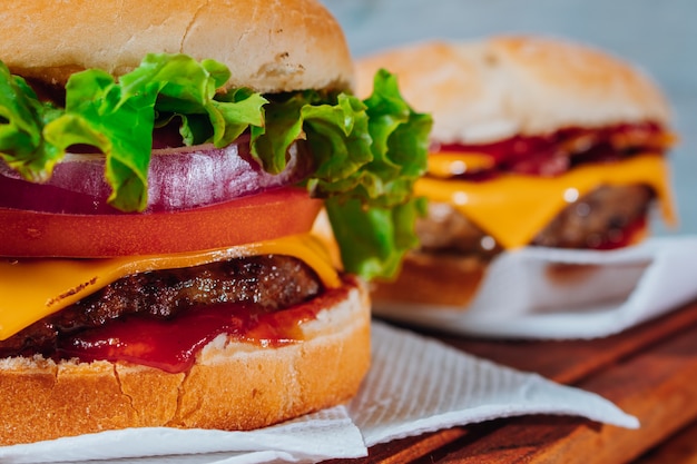 Deliciosos hambúrgueres com bacon e queijo cheddar e com alface, tomate e cebola roxa e bacon no pão caseiro e ketchup sobre uma superfície de madeira e fundo rústico. foco no primeiro hambúrguer.