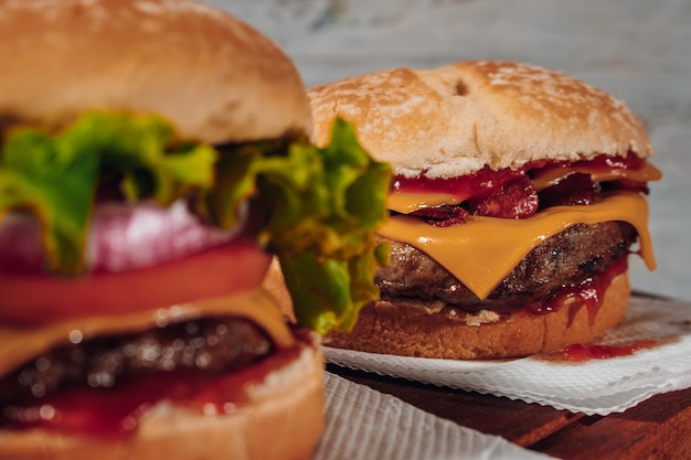 Deliciosos hambúrgueres com bacon e queijo cheddar e com alface, tomate e cebola roxa e bacon no pão caseiro e ketchup sobre uma superfície de madeira e fundo rústico. Foco no segundo hambúrguer.