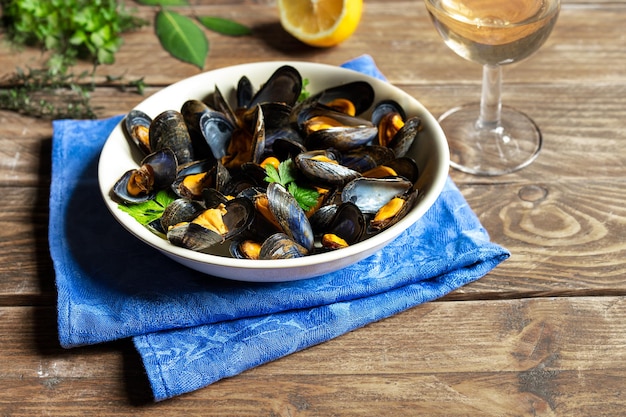 Deliciosos mejillones al vapor frescos en salsa de vino blanco en un plato blanco, perejil de limón y una copa de vino en una mesa de madera