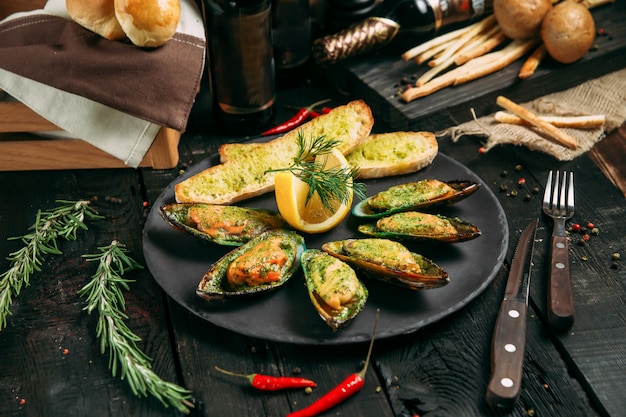 Deliciosos mejillones cocidos con salsa, pan de limón y ajo y mantequilla en un plato negro sobre el fondo de madera oscura