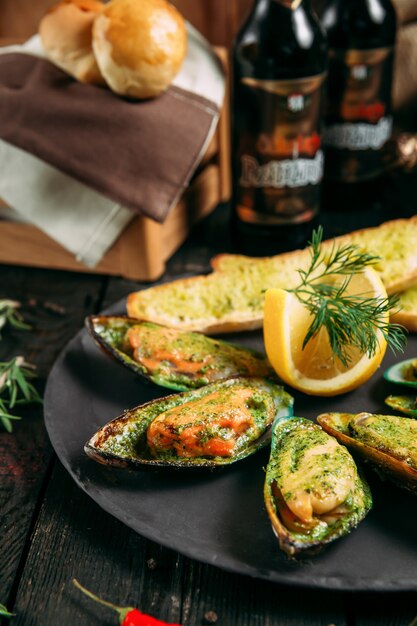 Deliciosos mejillones cocidos con salsa, pan de limón y ajo y mantequilla en un plato negro sobre el fondo de madera oscura