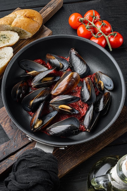 Deliciosos mejillones de mariscos cocidos con salsa de tomate con baguette, en una sartén de hierro, sobre fondo de mesa de madera negra