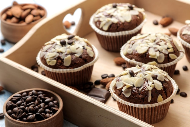 Deliciosos muffins de chocolate dulce, con pétalos de almendra en una bandeja de madera junto a granos de café en una mesa de madera azul.