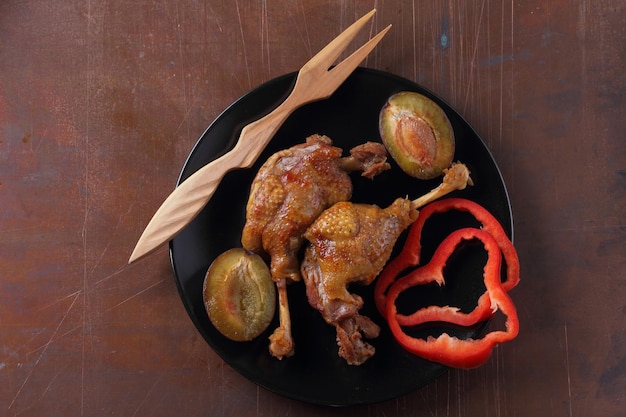 Deliciosos muslos de pato con corteza dorada y primer plano de un tenedor de madera Dos muslos de pato al horno con pimiento rojo y ciruelas en un plato negro sobre un fondo oscuro