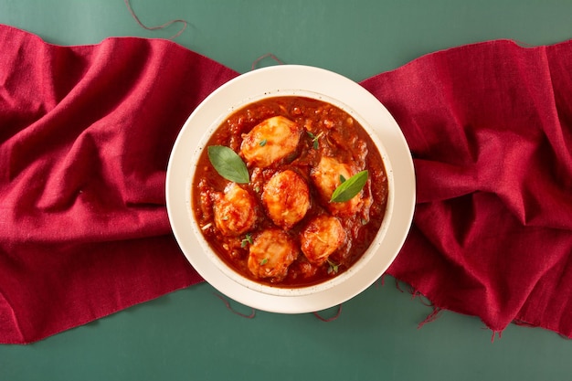 Deliciosos ñoquis con salsa roja y albahaca servidos en un tazón de cerámica hecho a mano con textura blanca en un fondo verde decorado con tela roja cruzando el marco en la vista superior