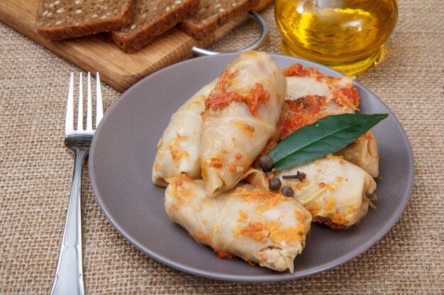 Deliciosos rollos de repollo caseros rellenos de arroz y carne en un plato con un tenedor, una botella de aceite de girasol y pan en una tabla de cortar de madera sobre una mesa con cilicio.