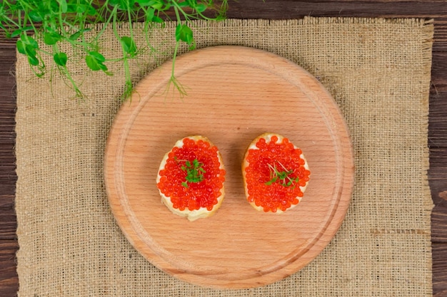 Deliciosos sándwiches con caviar rojo y mantequilla en una tabla de madera sobre un fondo de hierbas y champiñones Vista desde arriba