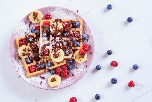 Deliciosos waffles belgas com mirtilos, framboesas, bananas e açúcar em pó são preenchidos com chocolate líquido. vista do topo.