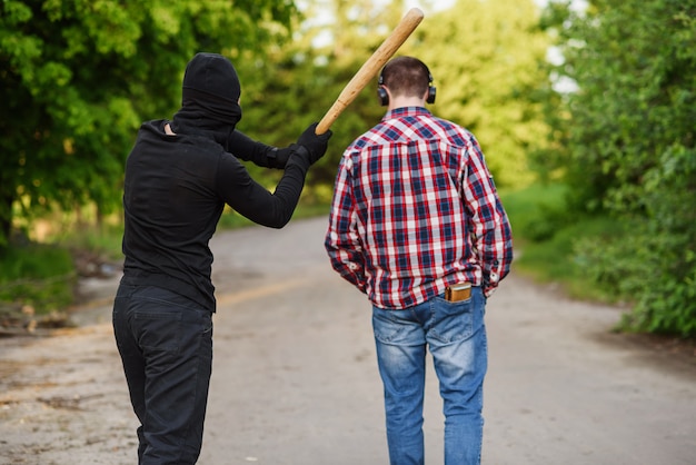 Un delincuente vestido de negro con un bate de béisbol en sus manos ataca a un hombre. Robar en la calle durante el día.