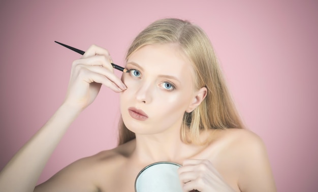 Delineador de ojos atractiva mujer rubia aplicando maquillaje mujer sostiene espejo y usando luz de lápiz cosmético