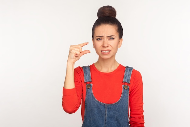 Demasiado pequeña. Retrato de una chica insatisfecha con moño de pelo en overoles de mezclilla mostrando un pequeño gesto y mirando con una expresión escéptica decepcionada. Foto de estudio interior aislado sobre fondo blanco.