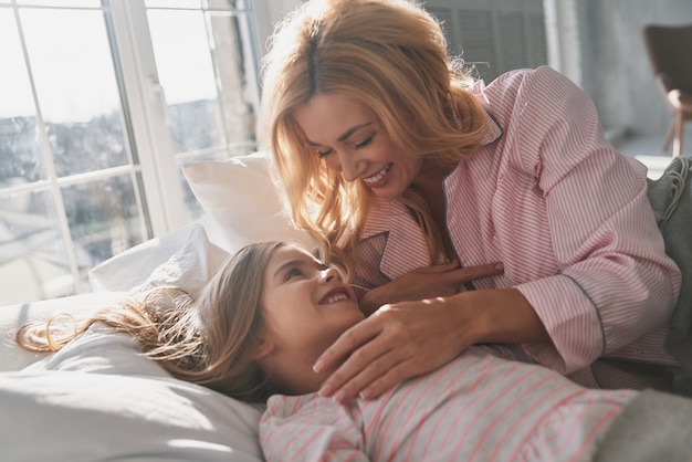 Demasiado perezoso para despertar. Hermosa joven madre despertando a su linda hijita y sonriendo mientras está acostada en la cama en casa