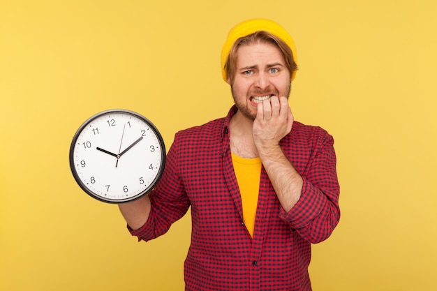 Demasiado tarde. Retrato de un hipster impaciente y ansioso con pantalones a cuadros sosteniendo un gran reloj y mordiéndose las uñas, nervioso por la reunión retrasada o la fecha límite, necesita darse prisa. tiro del estudio aislado en amarillo