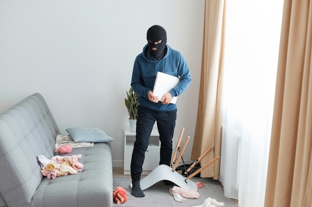 Dengerous mann in einer schwarzen maske und einem blauen kapuzenpulli betritt das haus eines anderen, um die besitzer auszurauben, die die wohnung aufbrechen, weil sie jemand anderem eigentum gestohlen haben, das einen laptop hält und weggeht