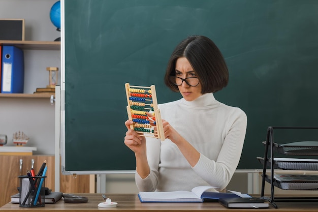 denkende junge Lehrerin, die eine Brille trägt und Abakus betrachtet, der am Schreibtisch mit Schulwerkzeugen im Klassenzimmer sitzt