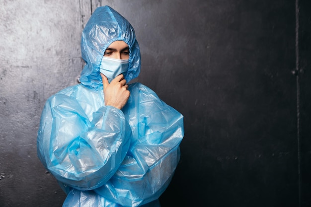 Denkender Arzt, der in der Nähe einer Wand steht und Schutzanzug und Gesichtsmaske trägt