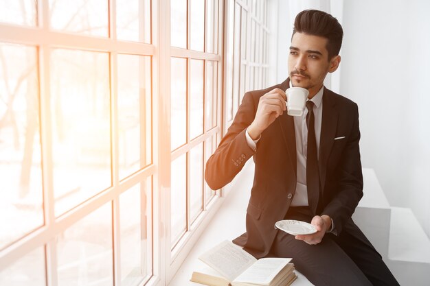 Denkender Geschäftsmann im schwarzen Anzug sitzt auf der Fensterbank und trinkt Tee, Sonnenlicht. Erfolgreiches Geschäftskonzept