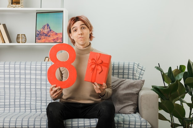 Denkender gutaussehender Kerl am glücklichen Frauentag, der die Nummer acht mit Geschenk auf dem Sofa im Wohnzimmer hält