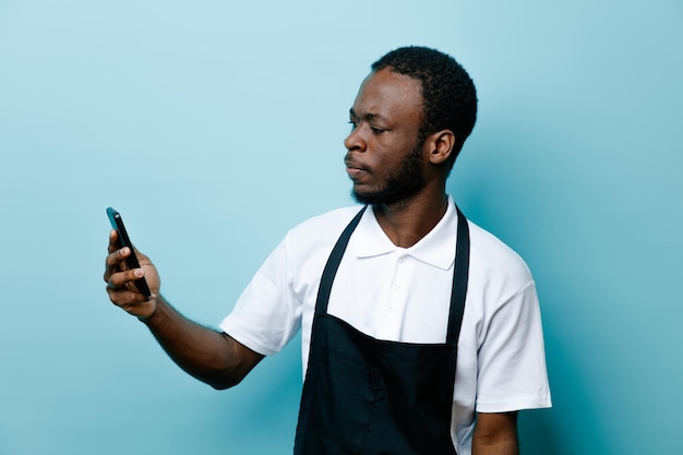 Denkender junger afrikanisch-amerikanischer Friseur in Uniform, der das Telefon isoliert auf blauem Hintergrund hält und betrachtet