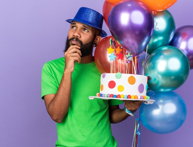 Denkender junger afroamerikanischer Mann mit Partyhut, der Luftballons mit Kuchen hält, packte das Kinn isoliert auf blauer Wand