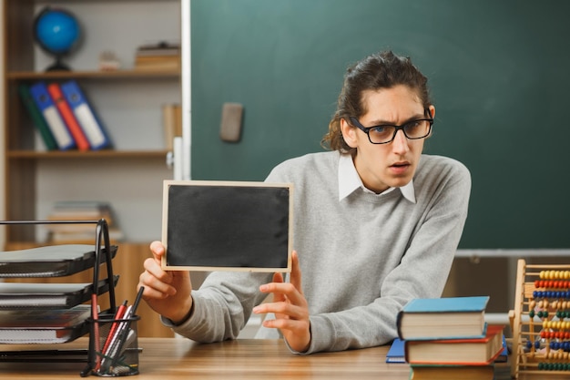 denkender junger männlicher lehrer mit brille, der am schreibtisch sitzt und eine mini-tafel mit schulwerkzeugen im klassenzimmer hält