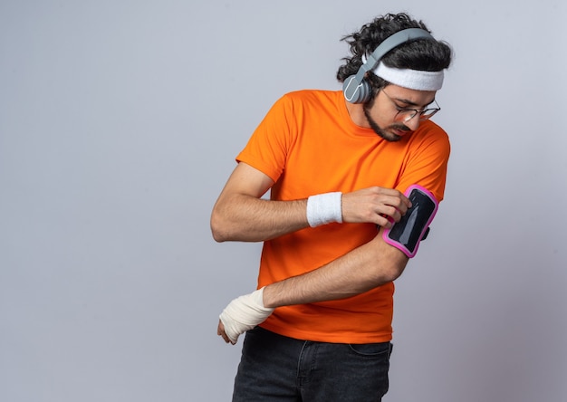 Denkender junger sportlicher mann mit stirnband mit armband und kopfhörern mit telefonarmband verletztes handgelenk mit verband umwickelt