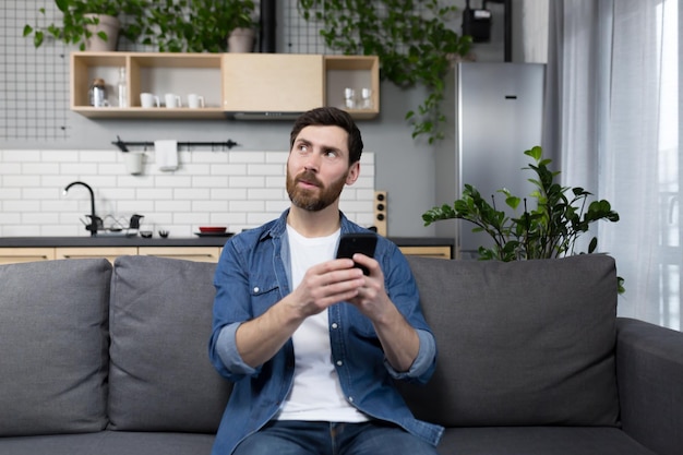 Denkender Mann schaut auf den Telefonbildschirm, der zu Hause auf dem Sofa sitzt