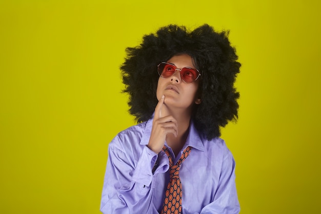 Denkendes Mädchen in Brille und Männerhemd und Krawatte mit afro lockiger Frisur auf gelber Wand