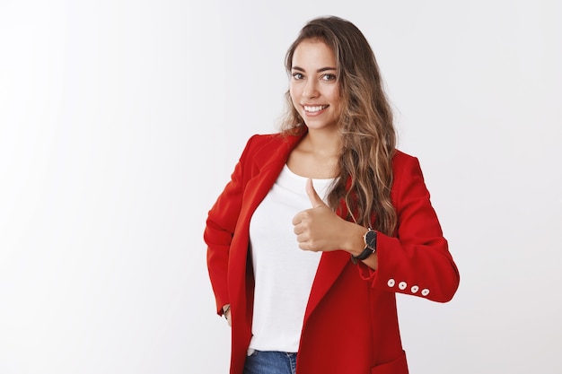 Foto dentes brancos alegres de mulher caucasiana de cabelos encaracolados sorrindo usando uma jaqueta vermelha da moda aparecendo o polegar como um gesto de aprovação, sorrindo em aprovação, parabenizando o amigo, ótima escolha, bom trabalho